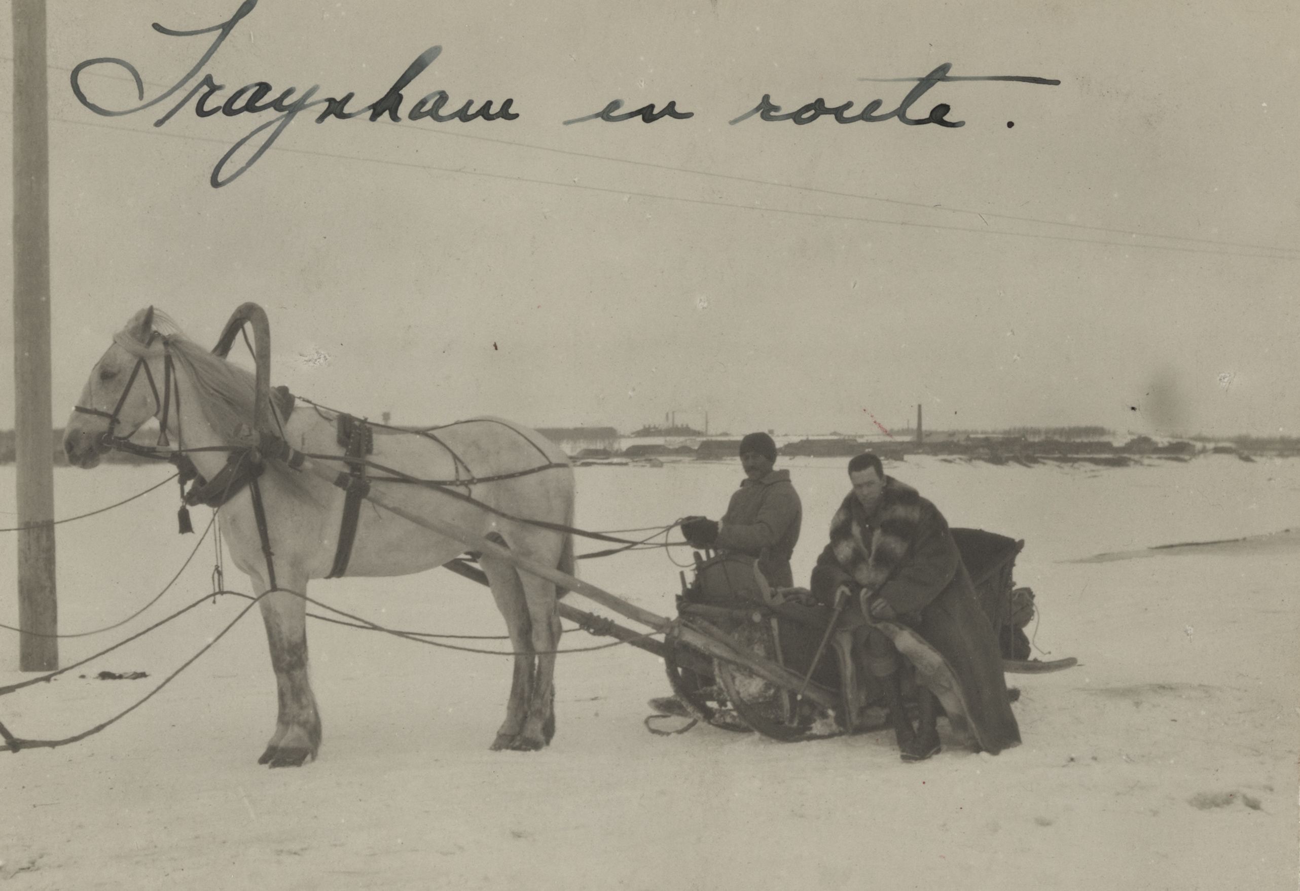Black and white photograph of Floyd Traynham in a heavy winter coat seated on a wooden sleigh next to sleigh driver who holds the reins for a white horse which the sleigh is hitched to.