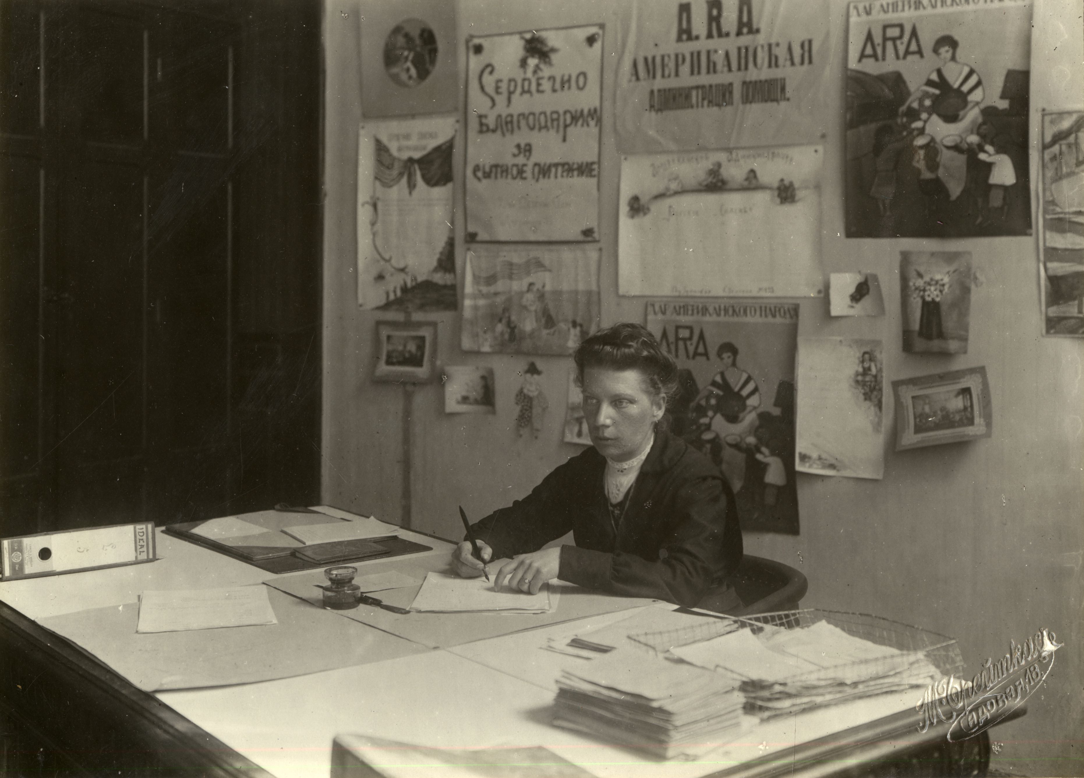 Black and white photograph of Lizzie Bach seated at her ARA office desk with several ARA posters on the wall behind her.