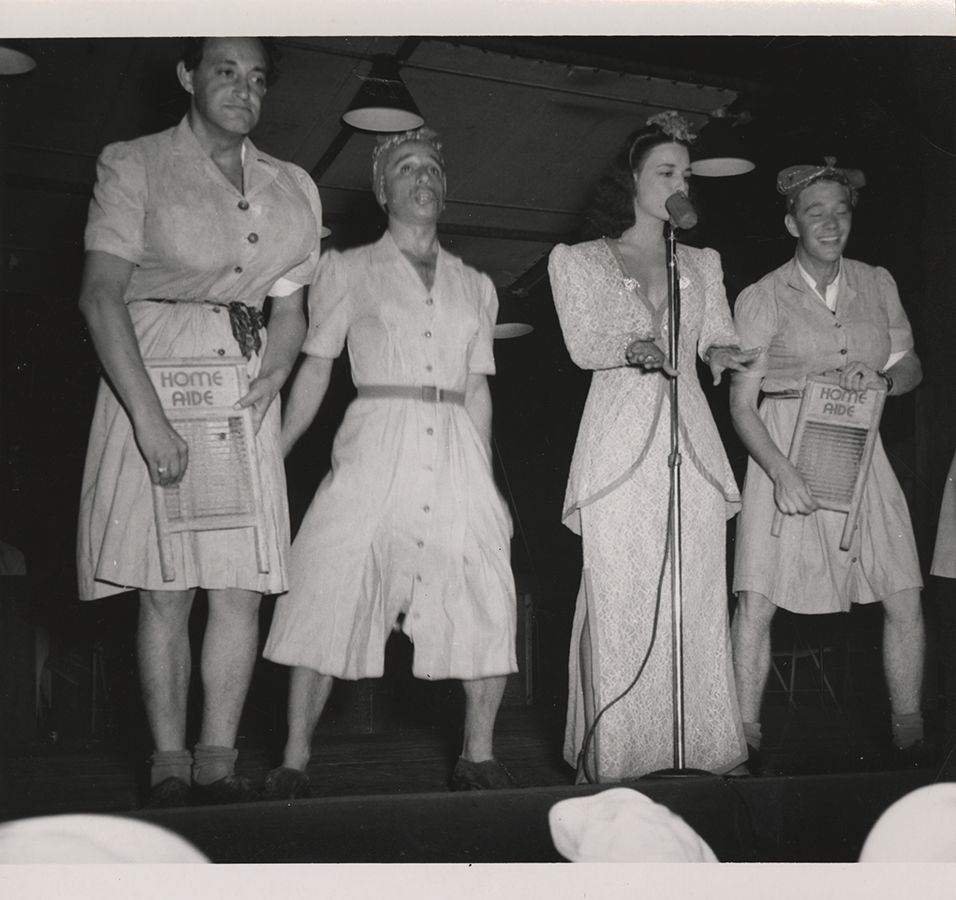 Black and white photograph of four USO performers on stage in New Caledonia, three men dressed as women and one woman at the microphone, all from the "Fun for Your Money" ensemble, 1945.