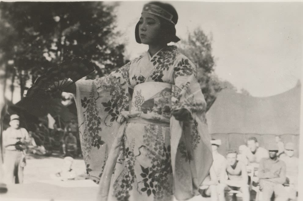 Black and white photograph of a Japanese woman in kimono performing a traditional dance with a fan in front of American troops, Okinawa, 1945.