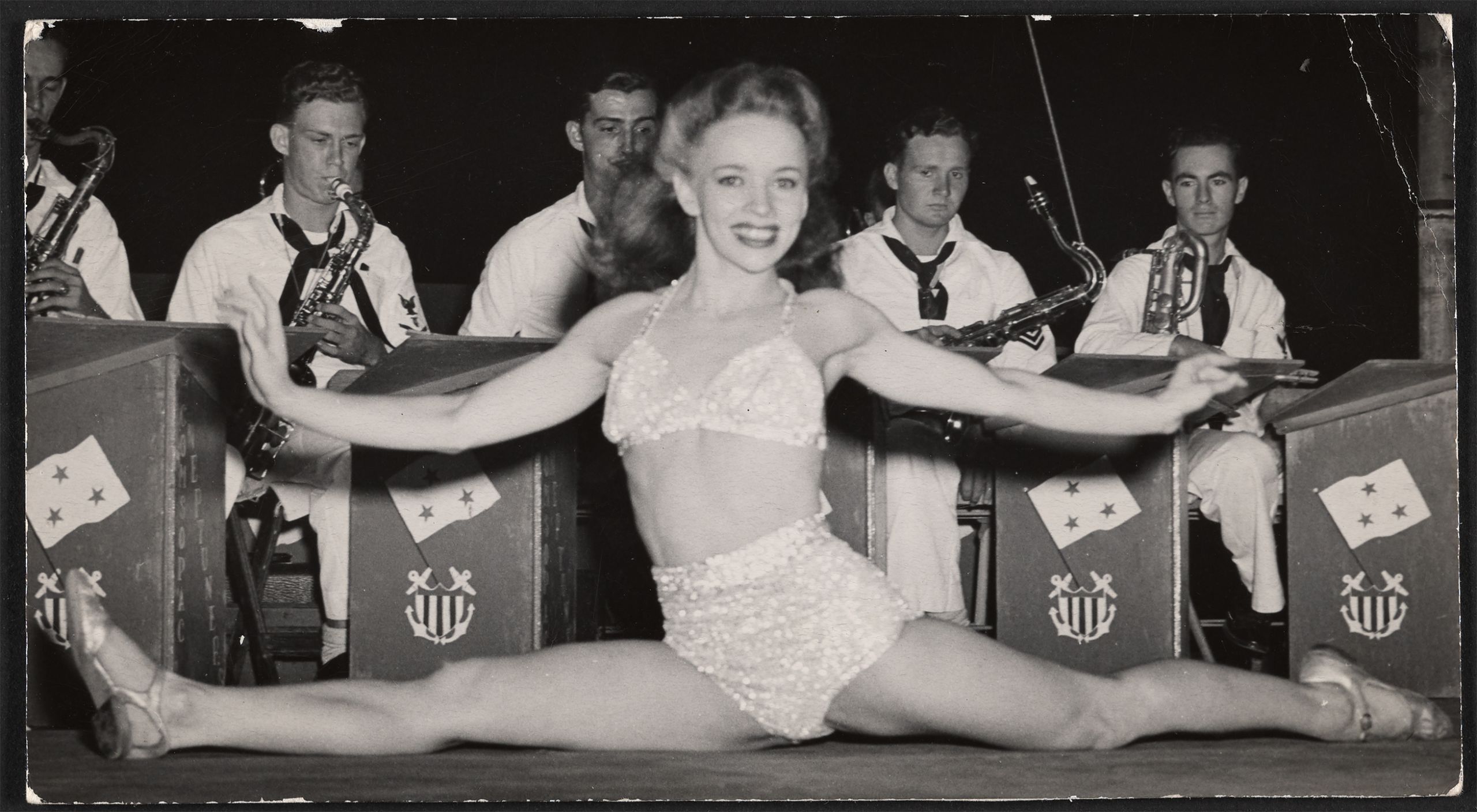 Black and white photograph of a woman doing a split in front members of the Neptunes playing their saxophones, 1945.