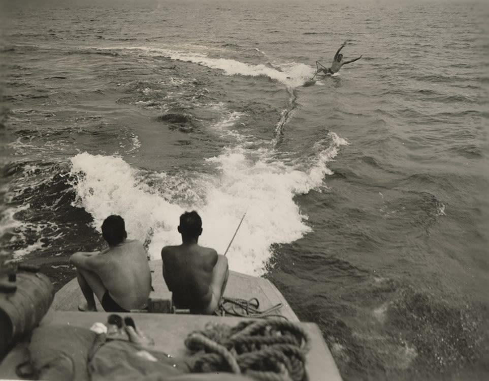 Black and white photograph of a Seabee falling over while water skiing, while two others watch on the back of a motor boat, 1945.