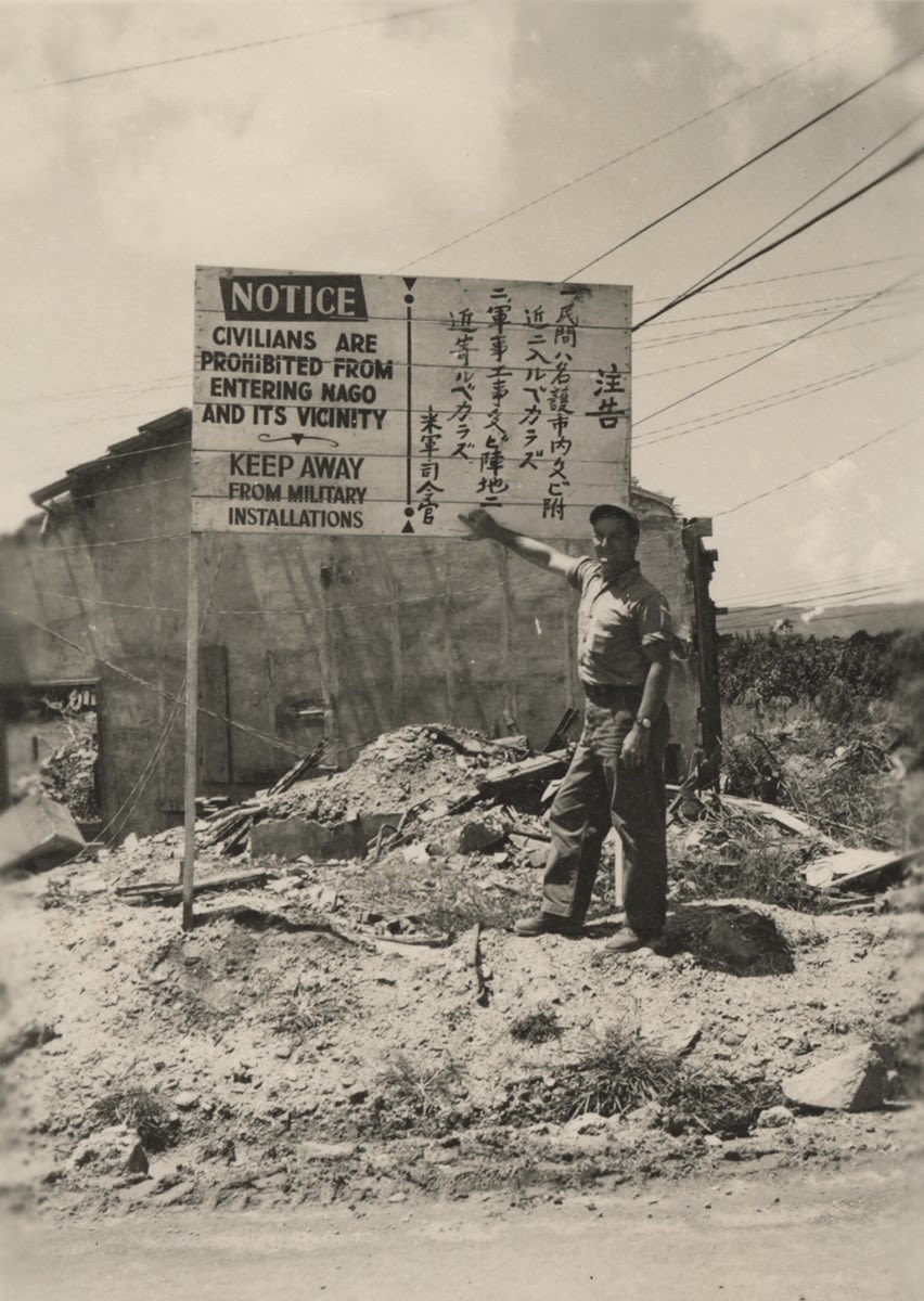 Black and white photograph of a Seabee pointing to a sign in both English and Japanese stating that Civilians are prohibited from entering Nago and its vicinity, Okinawa, 1945.
