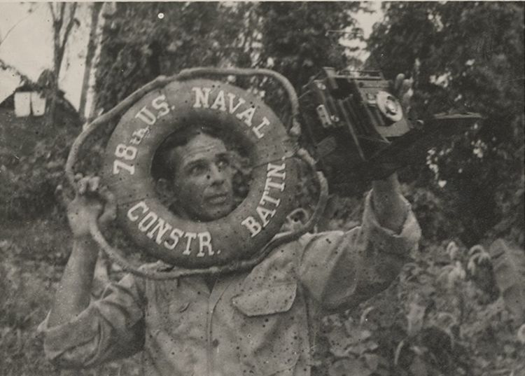Black and white photograph of photographer Edwin Keegan holding a lifesaver that has the text "78th US Naval Constr. Battn." written on it, and holding his camera in the other hand, no date.